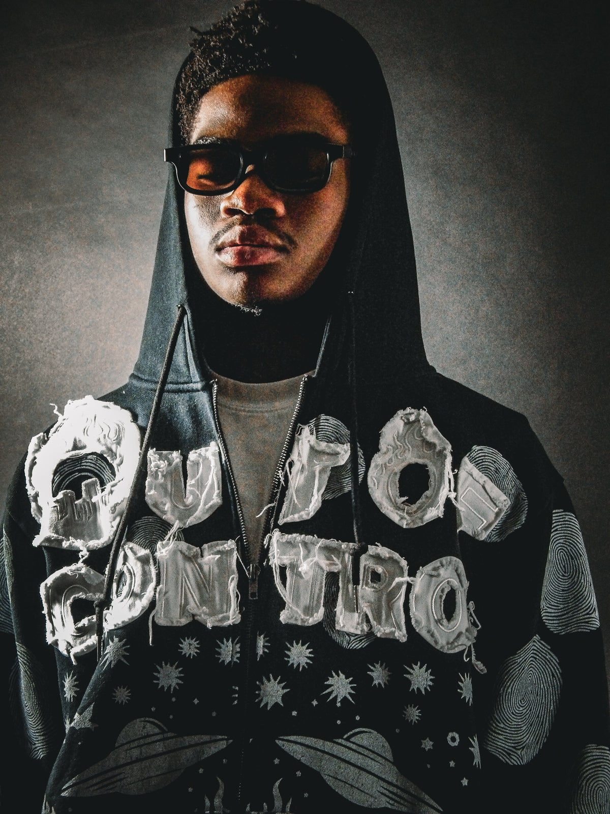 A person wearing a black hooded sweatshirt with large distressed white 'OUT OF CONTROL' lettering and dark abstract graphics, styled with black sunglasses against a dark gray studio background.
